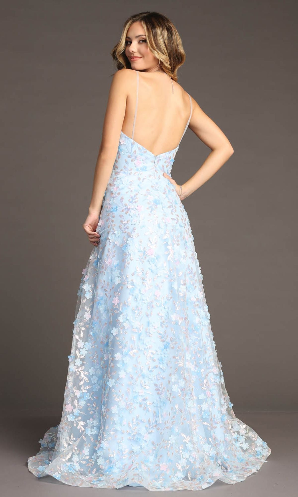 A woman wears the Chicas CHF3092 Long Light Blue Floral Prom Dress with delicate 3-D flowers and thin straps, standing and looking over her shoulder against a plain background.